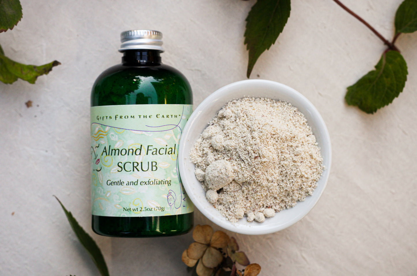 gifts from the earth almond facial scrub displayed next to the container surrounded by leaves